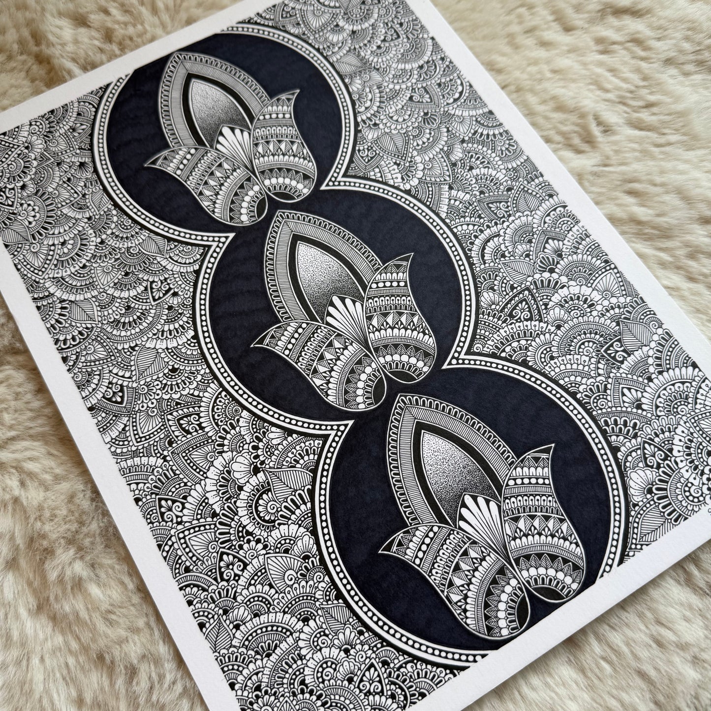 Hand-drawn zentangle by Saudamini Madra showing three lotus forms in black ink on Bristol paper, photographed on textured surface