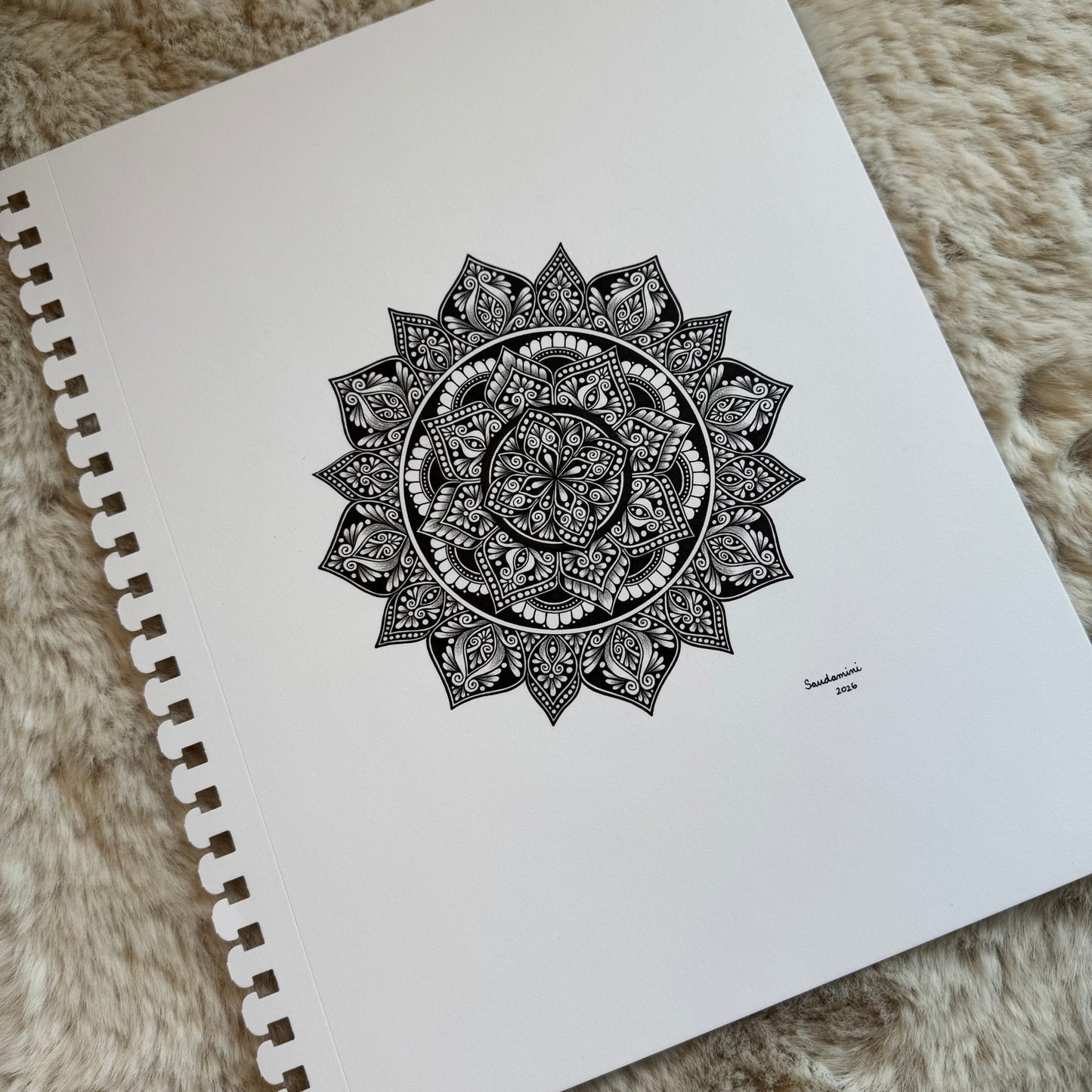 Hand-drawn mandala by Saudamini Madra in black ink on Bristol paper with geometric center and petal outer ring, photographed on textured surface