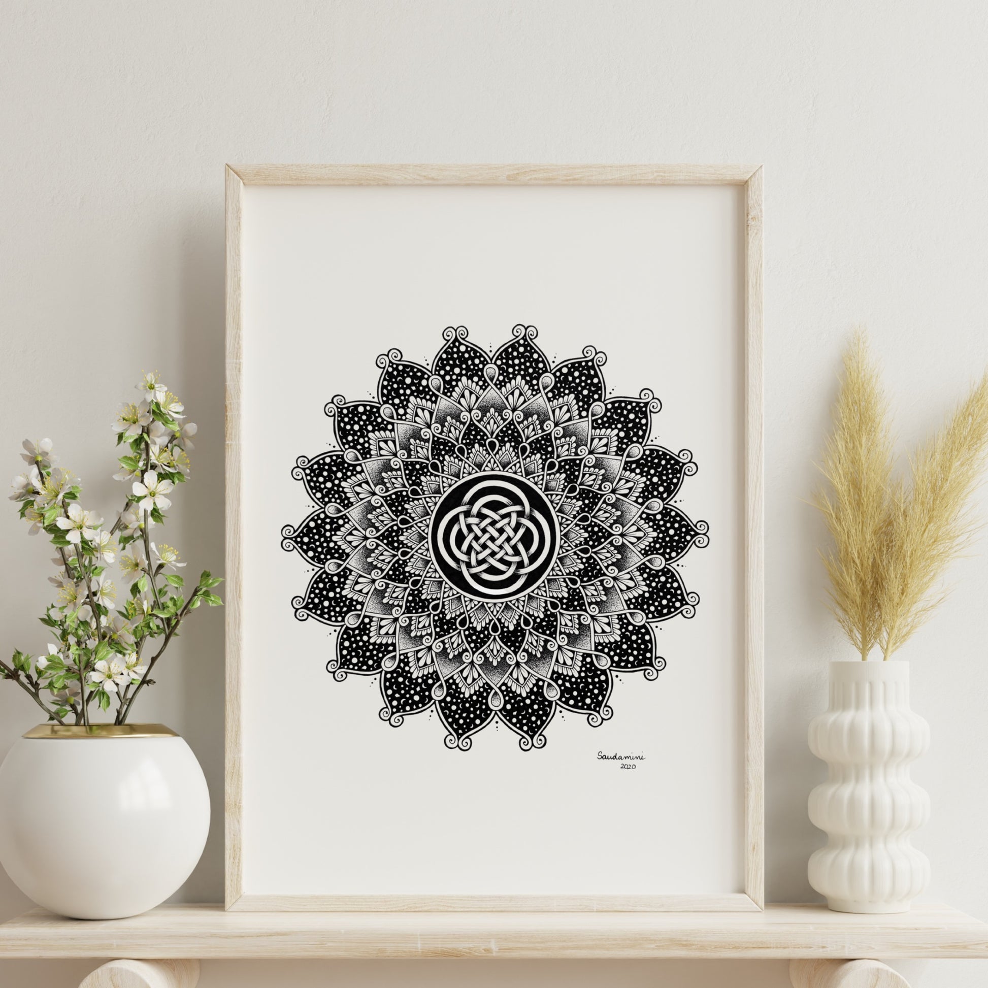 Composite mockup of The Knot That Holds the Night Mandala by Saudamini Madra, a circular mandala with Celtic knot center in black ink on Bristol paper, shown framed on a shelf