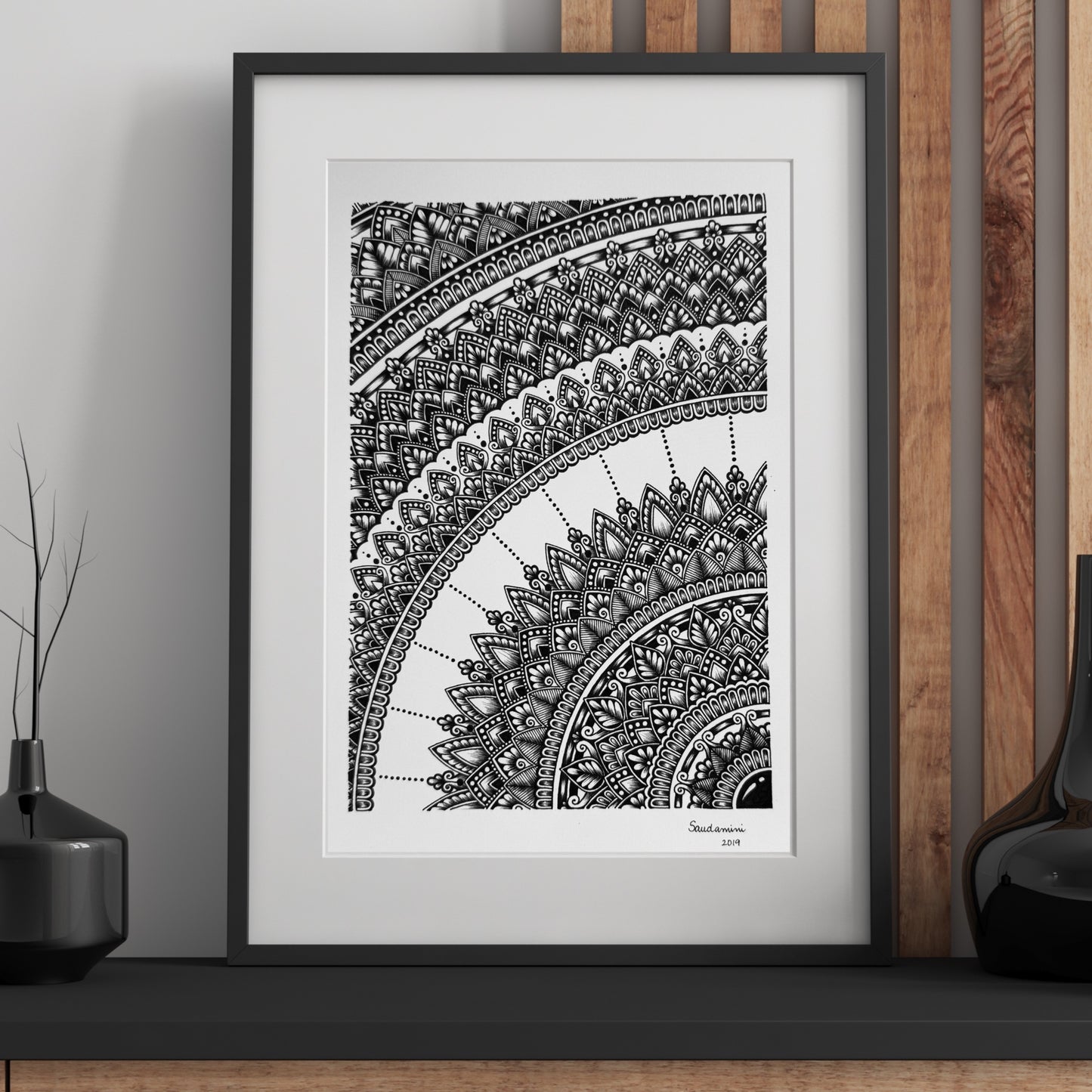 Composite mockup of The Memory of a Whole Mandala by Saudamini Madra, a cropped mandala arc in black ink on Bristol paper, shown framed against a wood-paneled wall