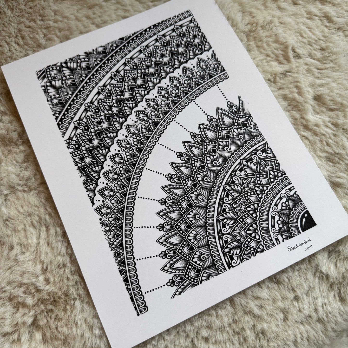 Hand-drawn mandala fragment by Saudamini Madra in black ink on Bristol paper, showing concentric arcs of fine patterning, photographed on textured surface