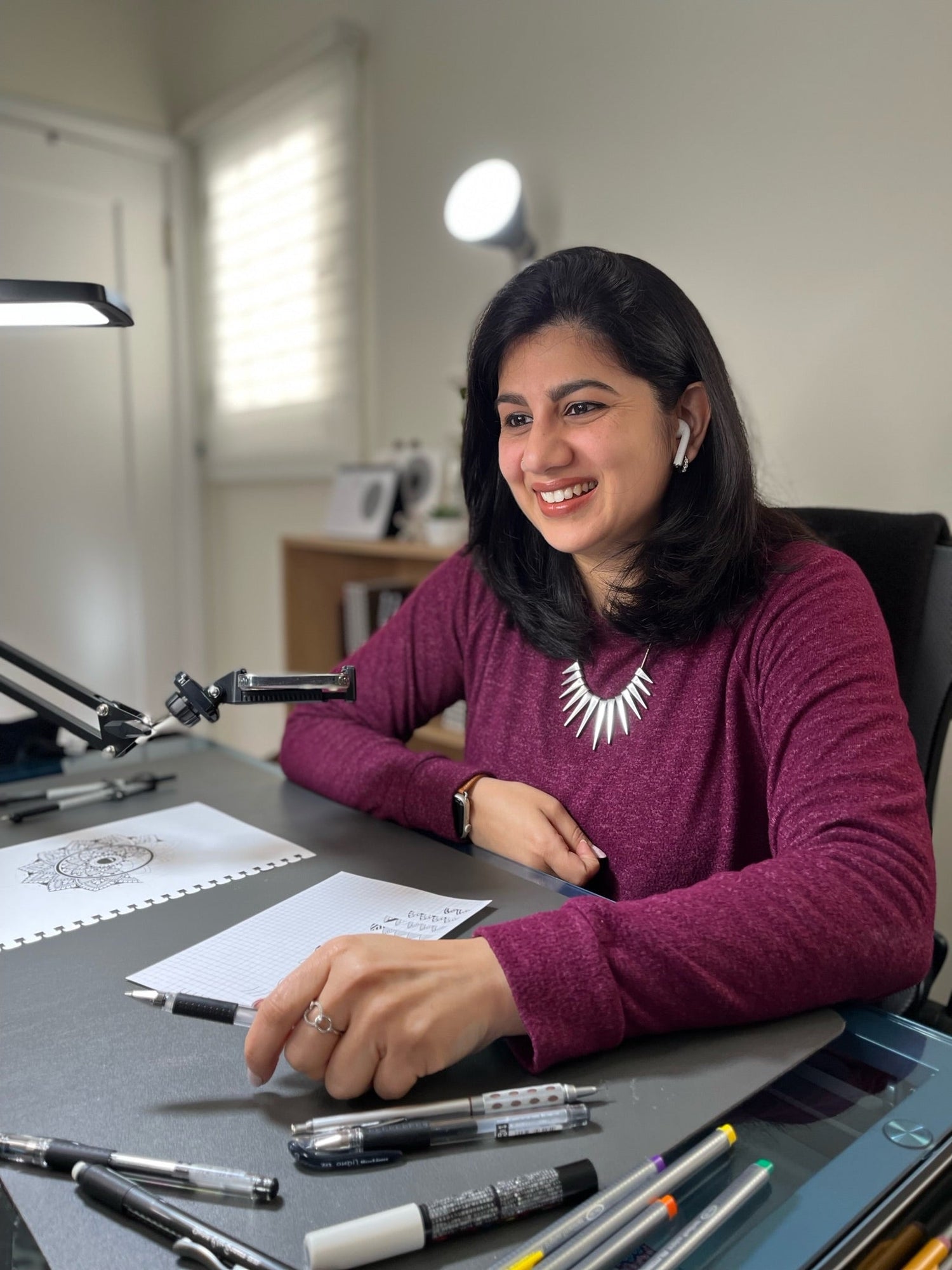 A smiling Saudamini Madra sitting at her studio with an in progress artwork on her desk